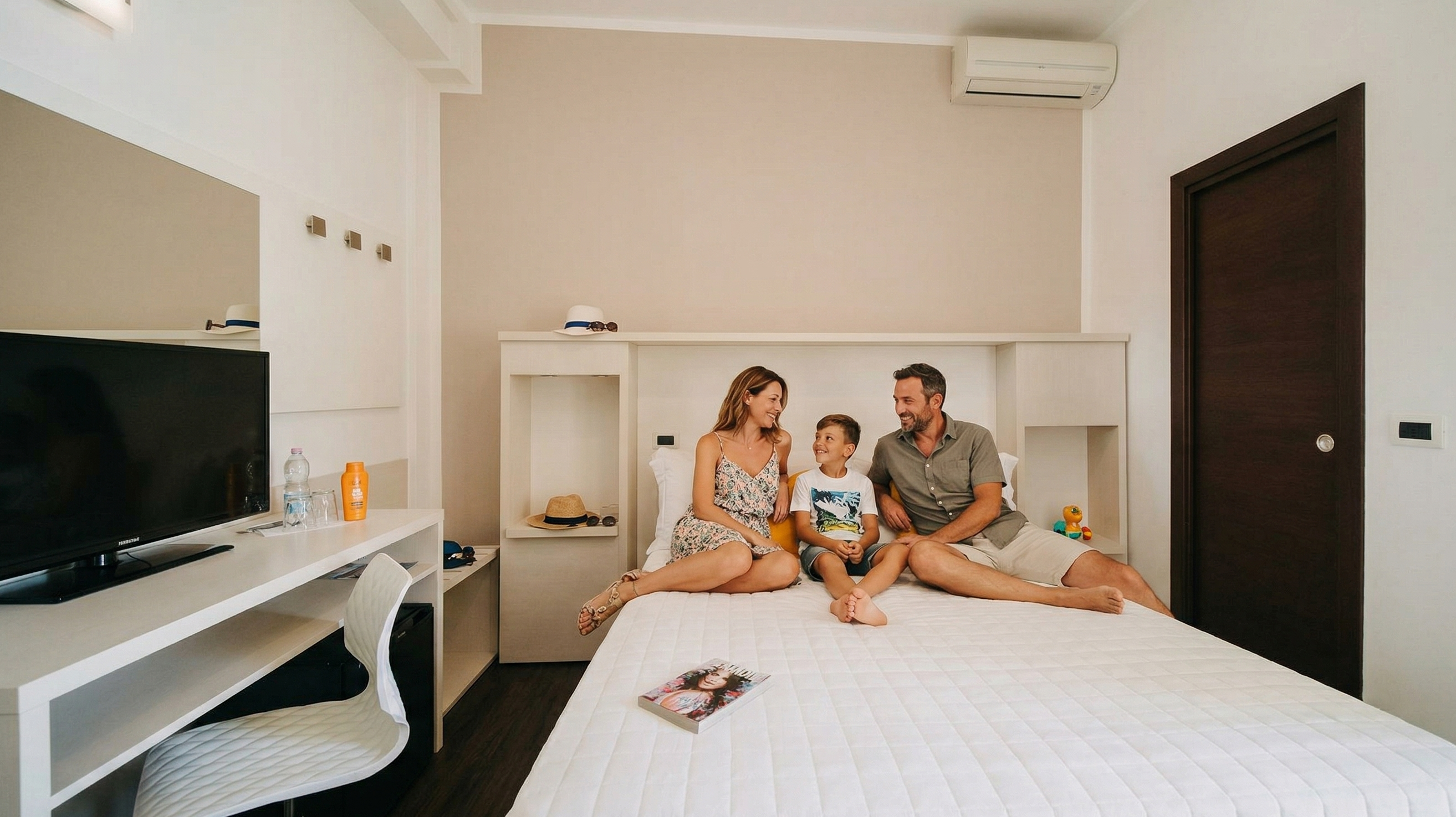 Una famiglia composta da un uomo e una donna di circa 40 anni e un bambino di 10 anni, vestiti con abiti estivi, seduti sul letto di una luminosa camera d'albergo moderna.
